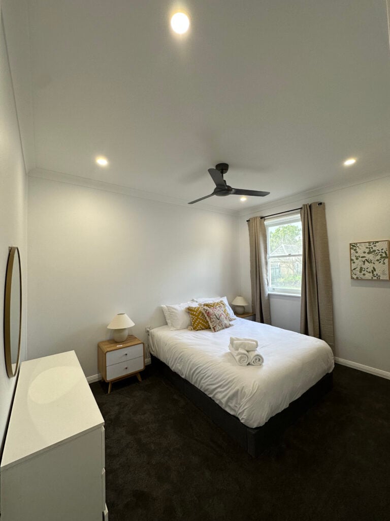 A brightly lit, modern bedroom featuring a double or queen-sized bed with white bedding.