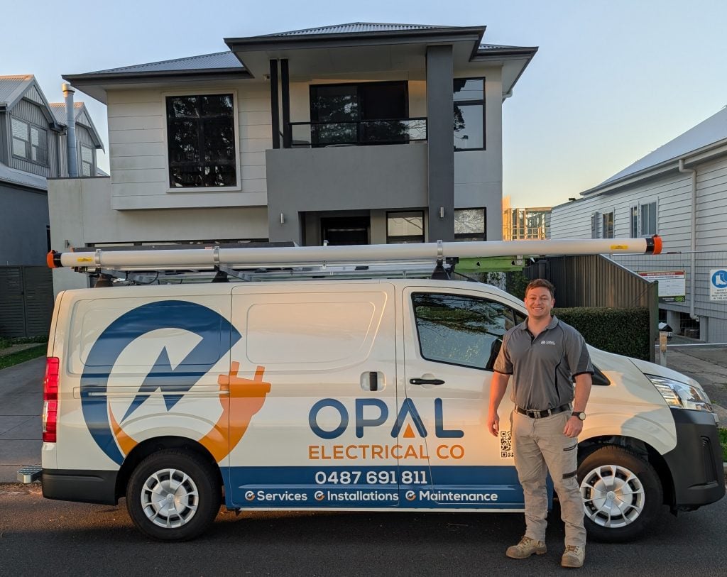 A smiling male electrician, wearing a grey polo shirt, tan work pants, and work boots, is standing next to a white service van.