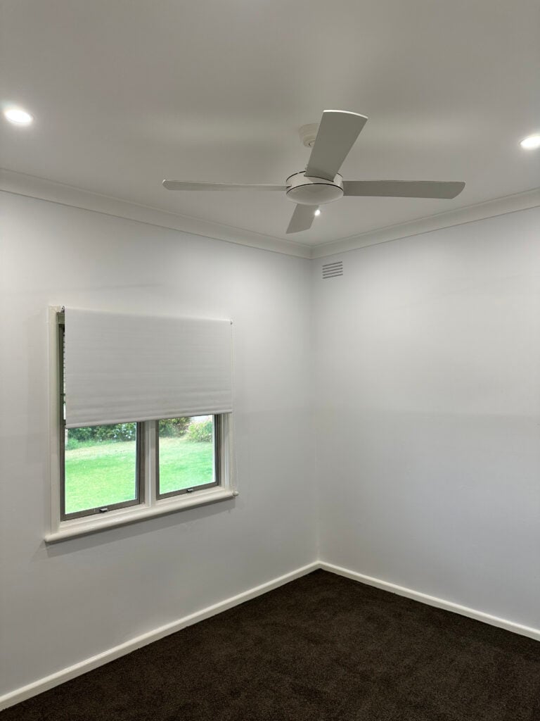 A corner view of a bright, empty room with plain white walls and dark brown carpet.