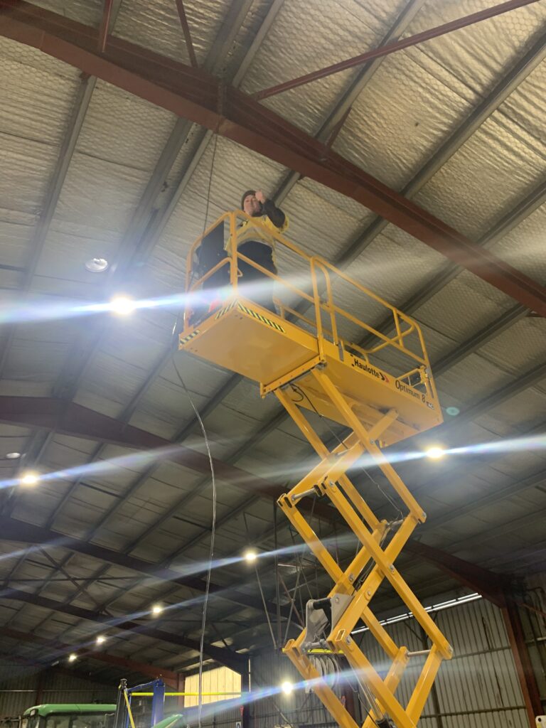 A male worker, wearing safety attire standing on the platform of a yellow scissor lift installing or repairing overhead lighting