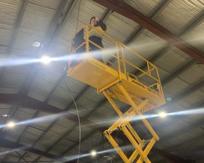 A male worker, wearing safety attire standing on the platform of a yellow scissor lift installing or repairing overhead lighting